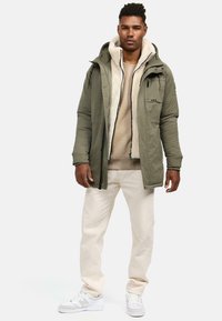Chaqueta acolchada verde con capucha forrada de pelo, sudadera beige debajo, pantalones crema y zapatillas blancas. Incluye bolsillos frontales y cremalleras.