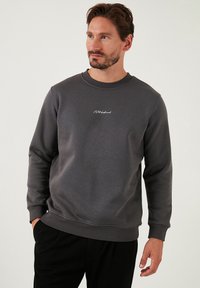 Šedá mikina s kulatým výstřihem, vyrobená z měkké látky, s malým bílým vyšitým logem na hrudi.