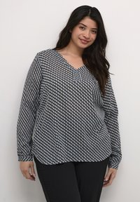 Long-sleeve, V-neck blouse featuring a black and white diamond pattern, semi-sheer fabric, and a relaxed fit. Paired with black pants.