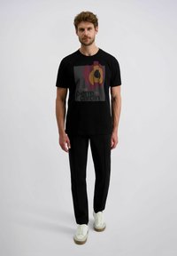 Mann steht in einem schwarzen, grafischen Pierre Cardin T-Shirt, schwarzen Hosen und weißen Sneakers mit braunen Sohlen vor einem einfarbigen, hellen Hintergrund.