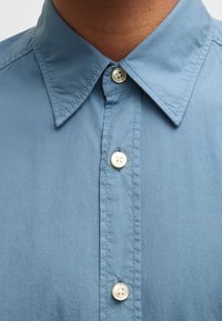 Camisa azul clara con un cuello clásico, con botones blancos y una tela suave, ligeramente texturizada. Diseño limpio con costuras visibles.