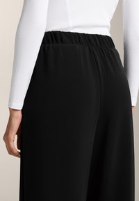 Fekete, rugalmas derekú nadrág sima textúrával, laza szabással. Egy formázott, fehér, hosszú ujjú felsővel viselve.