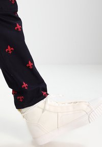 Bílé vysoké tenisky na šňůrky, kombinované s námořnickými kalhotami s červeným vzorem fleur-de-lis. Hladký povrch, neformální design.