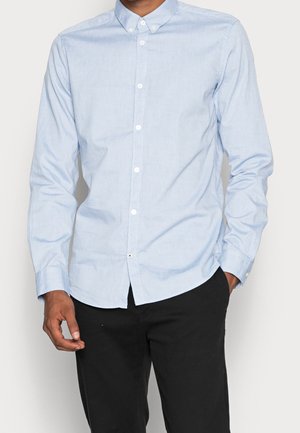 Mężczyzna w jasnoniebieskiej koszuli na guziki z długim rękawem i czarnych spodniach, stojący z jedną ręką w kieszeni na jednolitym tle.