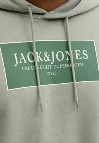 Jasnozielona bluza z kapturem z białą ramką logo zawierającą tekst "JACK & JONES" oraz "CREATIVE DEPT. COPENHAGEN" w kontrastowych kolorach.