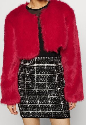 Kobieta w czerwonej kurtce z krótkim sztucznym futrem, nosząca czarną bluzkę i czarno-białą mini spódniczkę w kratkę z tkaniny tweed.