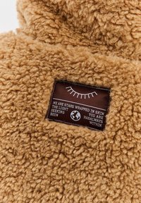 Hnědá fleece tkanina s přišitým obdélníkovým štítkem, na kterém je zobrazen měsíc řas, text a ikona zeměkoule.