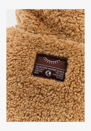 Braunes Fleece-Gewebe mit einem genähten rechteckigen Etikett, das einen Halbkreis von Wimpern, Text und ein Globus-Symbol zeigt.