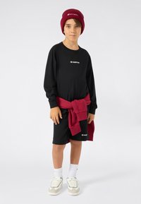 T-shirt à manches longues noir et shorts, pull bordeaux noué à la taille, chaussettes blanches, baskets avec accents bleus, arborant des logos Champion.