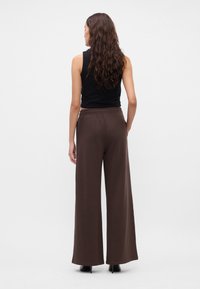 Καφέ παντελόνι φαρδιάς γραμμής με ελαστική μέση, κατασκευασμένο από μαλακό υλικό. Συνδυάζεται με μια εφαρμοστή μαύρη αμάνικη μπλούζα και μαύρες ψηλοτάκουνες γόβες.