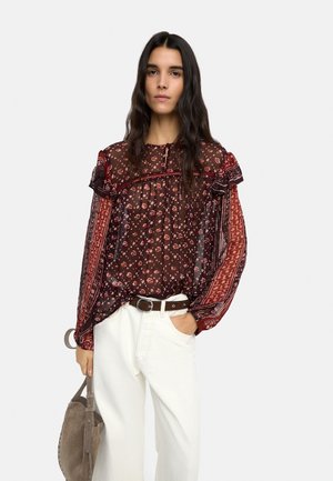 Kobieta o długich ciemnych włosach nosi przezroczystą bordową bluzkę z wzorem, białe spodnie z brązowym paskiem, trzyma szarobrązową zamszową torebkę na białym tle.