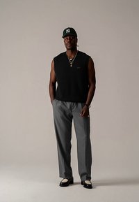 Moški, oblečen v črni brezrokavnik, sive hlače, črno-bele čevlje in zeleno baseball kapo, stoji z rokami v žepih na nevtralnem ozadju.