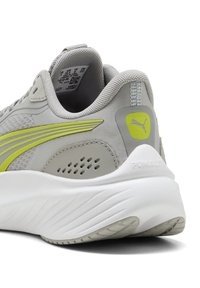 Zapato deportivo gris con detalles amarillos, parte superior de malla, cuello acolchado y suela blanca texturizada, con el logo de la marca en el talón y en el lateral.