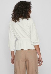 Blusa bianca, texturizzata, a maniche lunghe con vita cintrata e maniche a sbuffo. Abbinata a pantaloni beige, larghi e con tasche laterali.