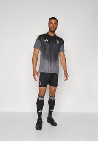 Moški nogometaš stoji oblečen v sivo-črni dres Juventusa, črne kratke hlače z roza črtami, črne nogavice do kolen in črne kopačke.