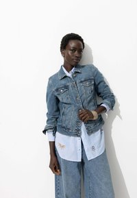 Veste en denim avec des boutons, des poches poitrine et une coupe ajustée, superposée sur une chemise rayée bleu clair avec des accents brodés.