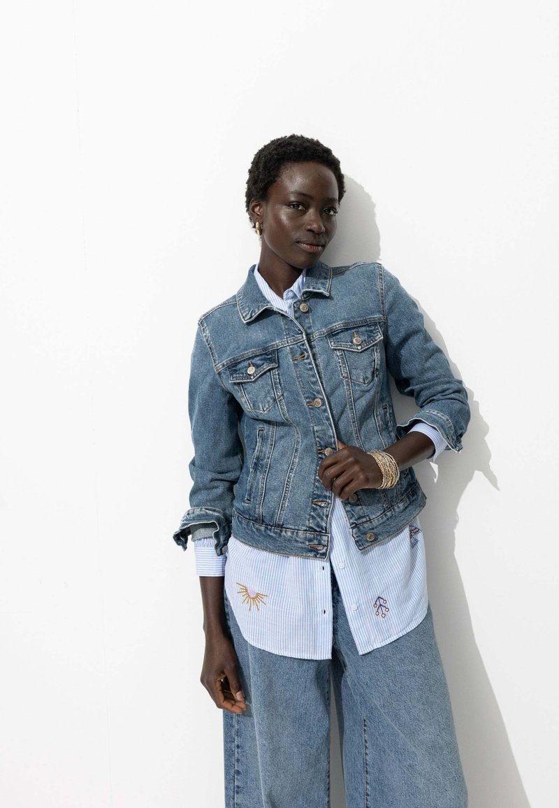 Veste en denim avec des boutons, des poches poitrine et une coupe ajustée, superposée sur une chemise rayée bleu clair avec des accents brodés.