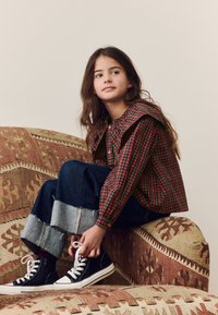 Chemise à carreaux rouge et verte avec un col froncé, accompagnée d'un jean en denim bleu avec des chevilles effilochées et des baskets hautes noires.