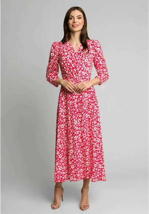 Kobieta stojąca w długiej czerwonej sukni z białym kwiatowym wzorem i bufkami na rękawach 3/4, w zestawieniu z beżowymi sandałami na wysokim obcasie.