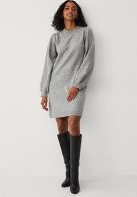 Grå strikket kjole med lange puffærmer, ribbede manchetter, let asymmetrisk kant, iført høje sorte læderstøvler.