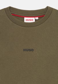 Žaliai olivinė palaidinė su iškilniu apvaliu kaklu. Papuošta juodu "HUGO" logotipu ant krūtinės ir raudona detale apykaklėje.