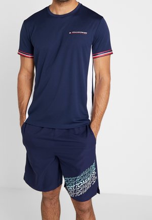 Mand iført navyblå kortærmet sportsskjorte med hvide sidepaneler og matchende navy shorts med lyseblå grafisk tekst.