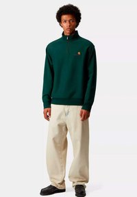 Sudadera de cremallera verde oscuro con cuello alto, que presenta un pequeño logotipo bordado. Combinada con pantalones holgados de color claro.