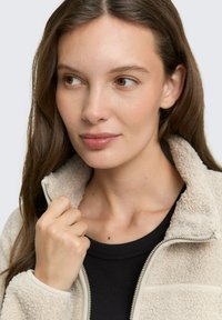 Veste en polaire beige avec un col zippé et des finitions crème. Le modèle tient le col d'une main, portant un t-shirt noir en dessous.