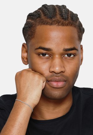 Gros plan sur une personne avec des cheveux tressés, portant un shirt noir. Un bracelet en argent est visible au poignet, avec une texture lisse et un design fin.