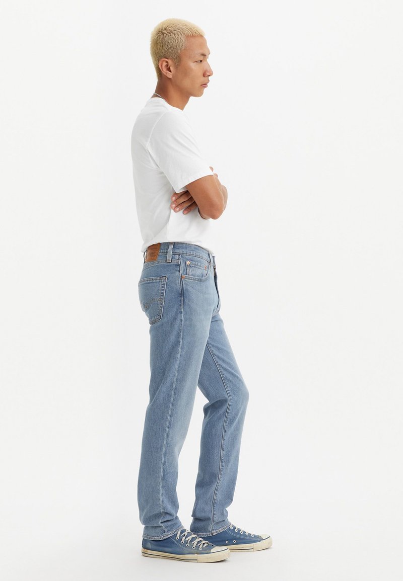 Jeans de mezclilla azul claro con corte recto, que cuentan con un parche de cuero marrón, cinco bolsillos y herrajes de metal plateado. Llevados con una camiseta blanca.