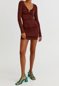 Braunes, langärmliges Bodycon-Kleid mit Twist-Detail und rundem Ausschnitt am Hals. Kombiniert mit grünen, sandalen mit blockigem Absatz und Riemchen.