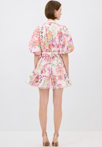 Bloemenjurk in crème met roze en groene patronen, met pofmouwen, een ceintuur in de taille en een ruches onderrand. Gladde stof met een kanten rand.