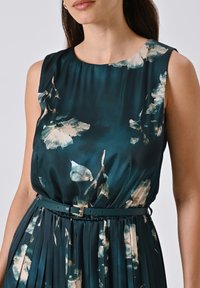 Ärmelloses türkisfarbenes Kleid mit floralem Muster, plissierter Rock und passendem Gürtel. Der Stoff hat eine satinähnliche Oberfläche, die eine glatte Textur erzeugt.