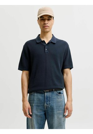 Férfi bézs sapkával, sötétkék rövid ujjú pólóinggel és világoskék farmerrel, egyszínű világos háttér előtt áll.