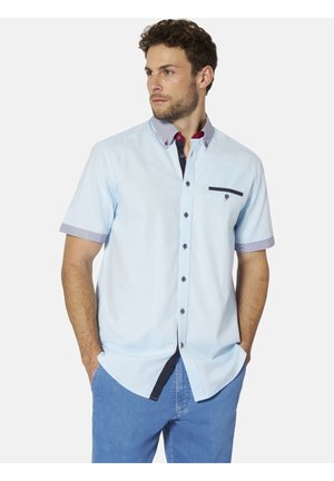 Mann mit kurzen lockigen Haaren, der ein hellblaues kurzärmeliges Hemd mit gemustertem Kragen, Knopfdetails und blaue Hosen trägt, Hände in den Taschen.