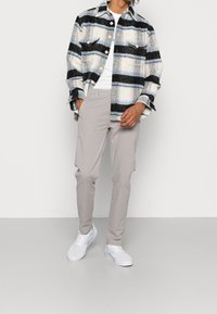 Chemise en flanelle avec un motif à carreaux noir, bleu et blanc sur fond crème ; portée avec un pantalon gris clair et des baskets blanches, debout sur un arrière-plan neutre.