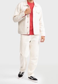 Veste en denim blanche portée sur un pull rouge en maille torsadée, associée à un pantalon blanc cassé et des baskets noires. Design simple avec des détails subtils.