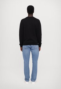 Man som står med ryggen mot kameran, iklädd en svart långärmad skjorta, ljusblå jeans och vita sneakers mot en enfärgad ljus bakgrund.