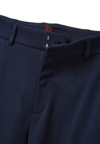 Pantaloni blu navy con una texture liscia, dotati di una vita piatta, due chiusure metalliche e dettagli di cucitura aggiuntivi.