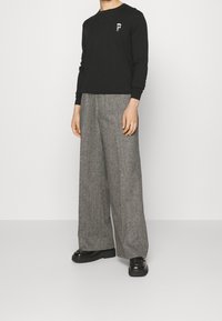 Personne portant un t-shirt à manches longues noir avec un petit graphique sur la poitrine, un large pantalon en tweed gris et des chaussures à plateforme noires, se tenant devant un fond uni.