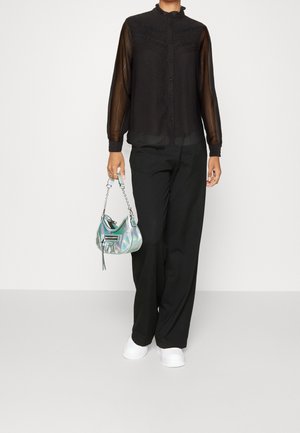 Personne portant un chemisier transparent noir à boutons, un pantalon large noir, des baskets blanches, tenant un petit sac irisé à bandoulière chaîne.