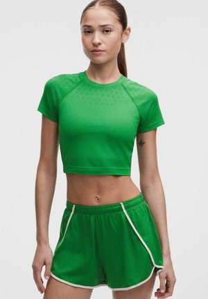 Joven mujer con una camiseta deportiva corta verde y pantalones cortos a juego con ribete blanco sobre un fondo liso.