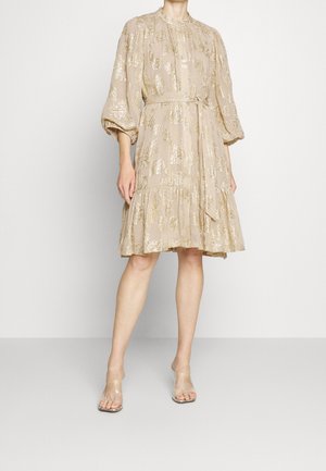 Beige knielange jurk met metalen gouden bloemmotieven, pofdriekwartmouwen, taillelint en transparante openhakken op een model.