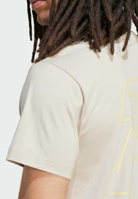 Camiseta de color beige claro, hecha de algodón, con mangas cortas y un patrón geométrico amarillo en la espalda. Textura suave, corte relajado.