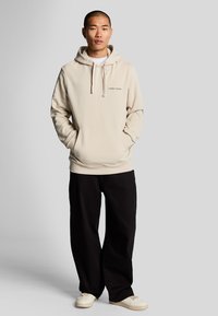 Sweat-shirt beige à capuche avec une poche avant et des cordons, associé à un pantalon noir ample et des chaussures claires. Détail de logo simple sur la poitrine.