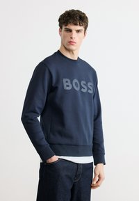 Νεαρός με μπούκλες στέκεται πρόσωπο με πρόσωπο, φορώντας μπλε σκούρο φούτερ με την επιγραφή «BOSS» και σκούρο τζιν παντελόνι.