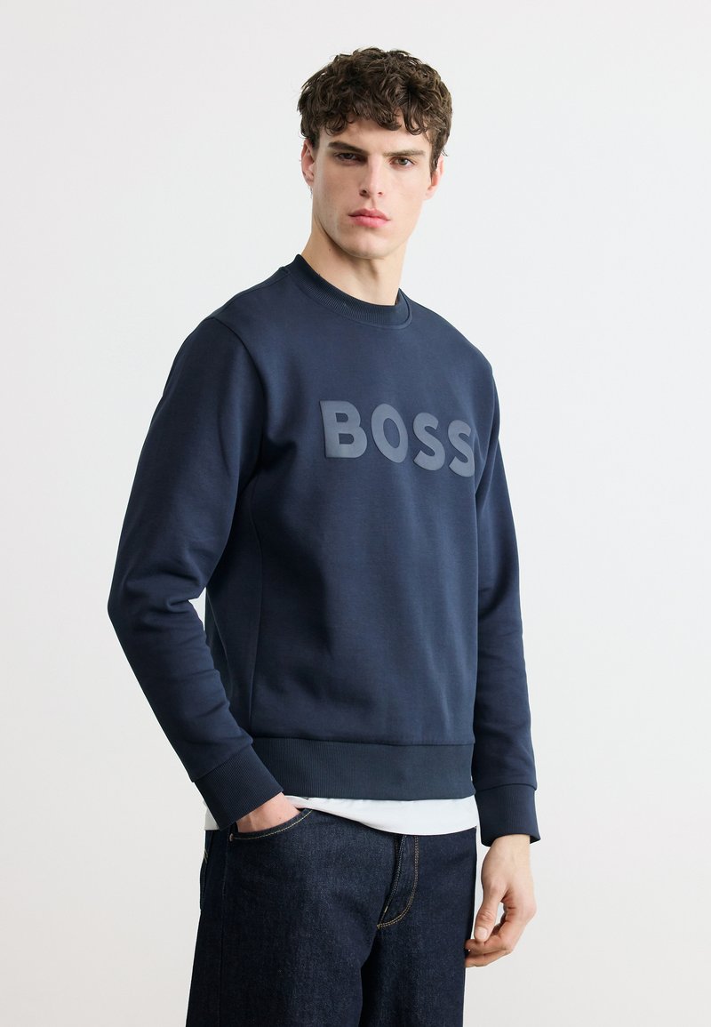 Νεαρός με μπούκλες στέκεται πρόσωπο με πρόσωπο, φορώντας μπλε σκούρο φούτερ με την επιγραφή «BOSS» και σκούρο τζιν παντελόνι.