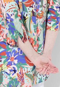 Top con motivo floreale caratterizzato da colori vivaci come verde, rosso, blu e rosa. Realizzato in tessuto leggero, con maniche a tre quarti e spacco laterale.