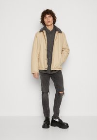 Chaqueta acolchada beige con un cuello forrado de piel gris, combinada con una camisa negra de botones y unos jeans negros rasgados, completada con unos zapatos negros voluminosos.