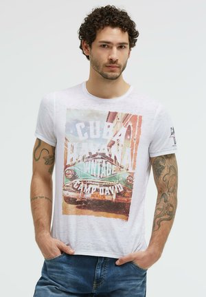 Man with curly hair wearing a white t-shirt with a vintage car and Cuba Havana print, standing with hands in pockets.